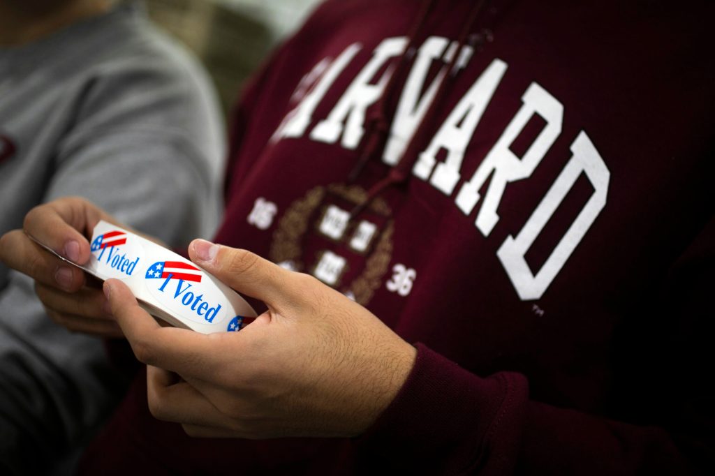 Person in a Harvard sweatshirt holds Vote stickers.