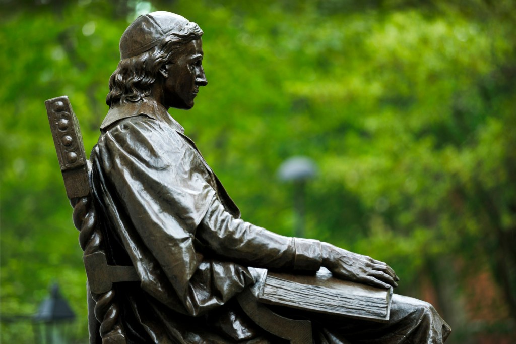 profile of the John Harvard statue against a backdrop of bright green leaves.