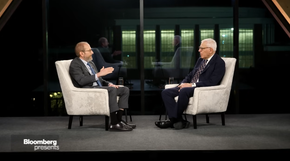 two men sitting in chairs on a stage talking.