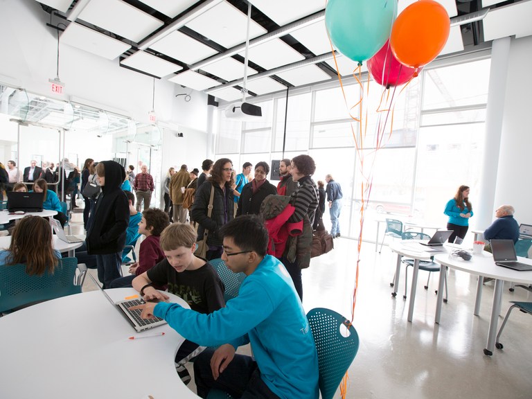 Two people sit at a table with a laptop, behind them is a group in a large sunny room with balloons.