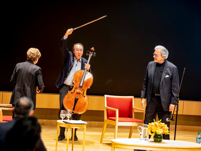 Men stand on a stage, one holding a cello