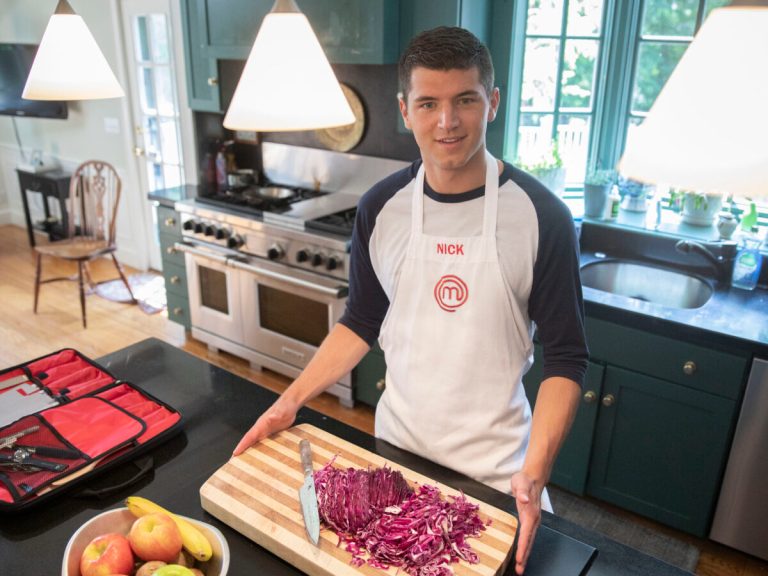 A student wearing an apron stands in a kitchen with a cutting board of purple cabbage in front of him