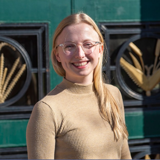 A young woman wearing a beige turtle neck poses outdoors in front of a green and gold wall.
