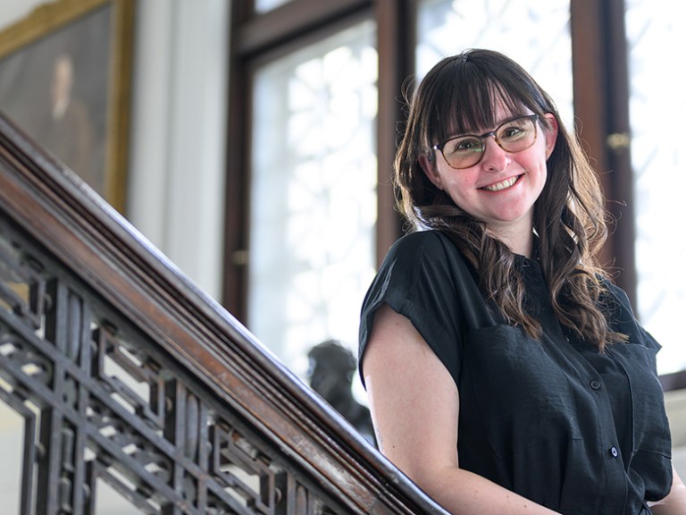 A smiling student stands on a staircase in front of a large window
