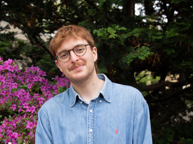 A young man poses in front of a bush with pink flowers