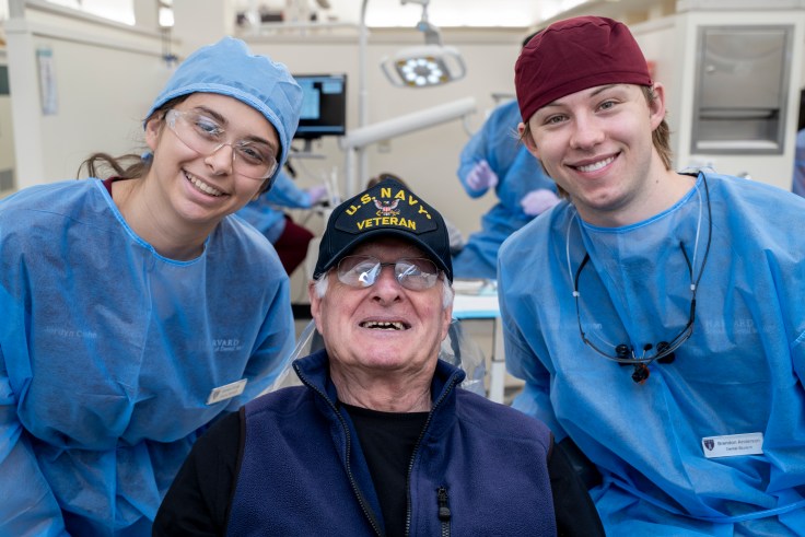 Two dental students wearing scrubs pose with an older man wearing a U.S. Navy hat.